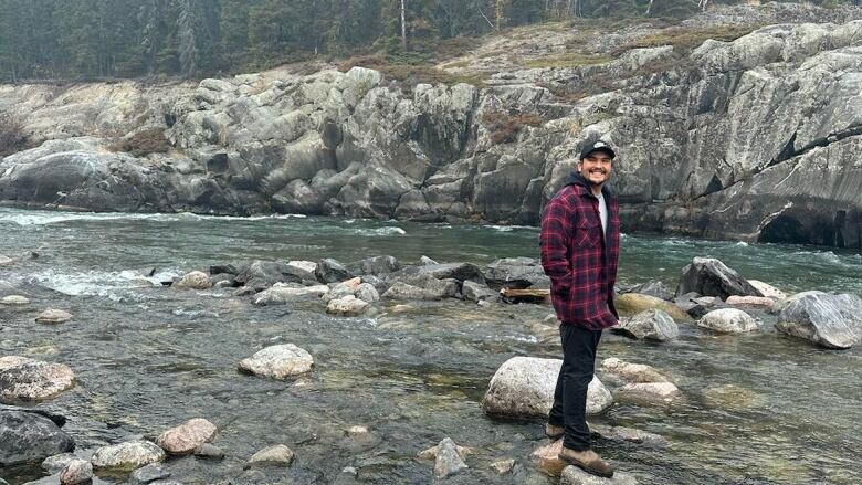 A young man stands in a river with trees behind him.