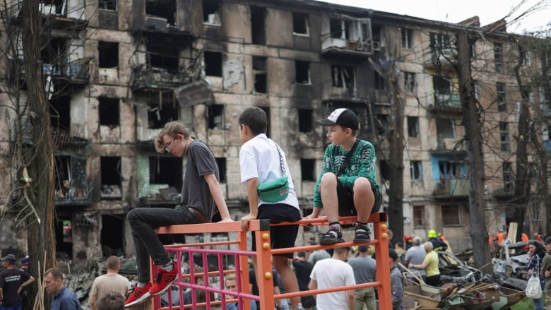 Children play on a structure, with a wrecked building in the background.