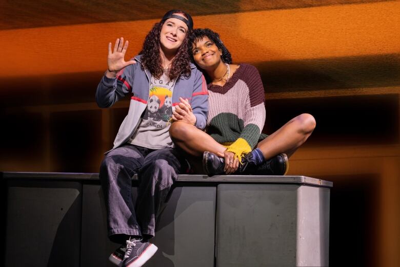 Two performers sit onstage. The person on the left is gesturing and smiling, while the person on the right leans their head on the other's shoulder. 