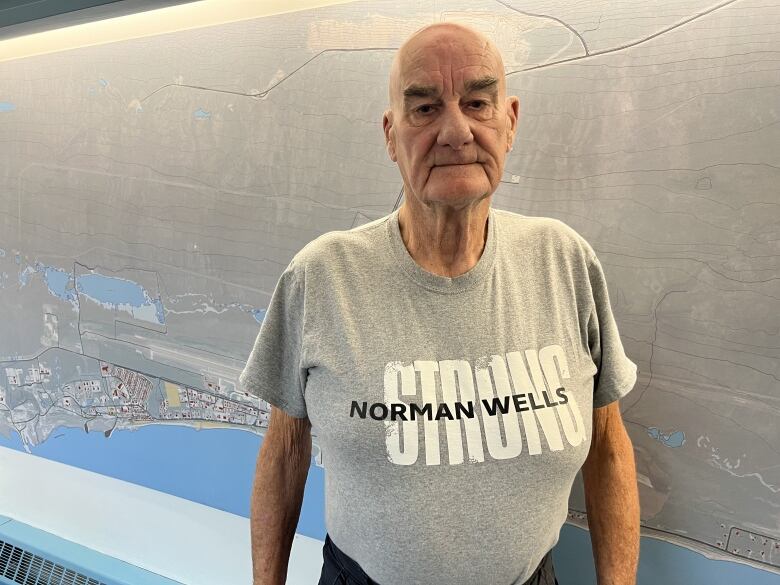 An elderly man in a t-shirt stands in front of a large map.