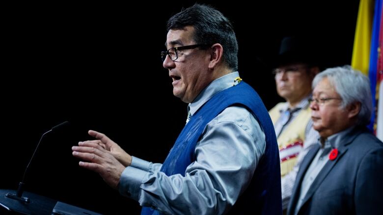 A man gestures while speaking at a podium, with two other men behind him.