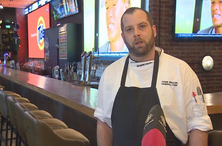 A white man wearing a chef's uniform speaks in front of a bar.