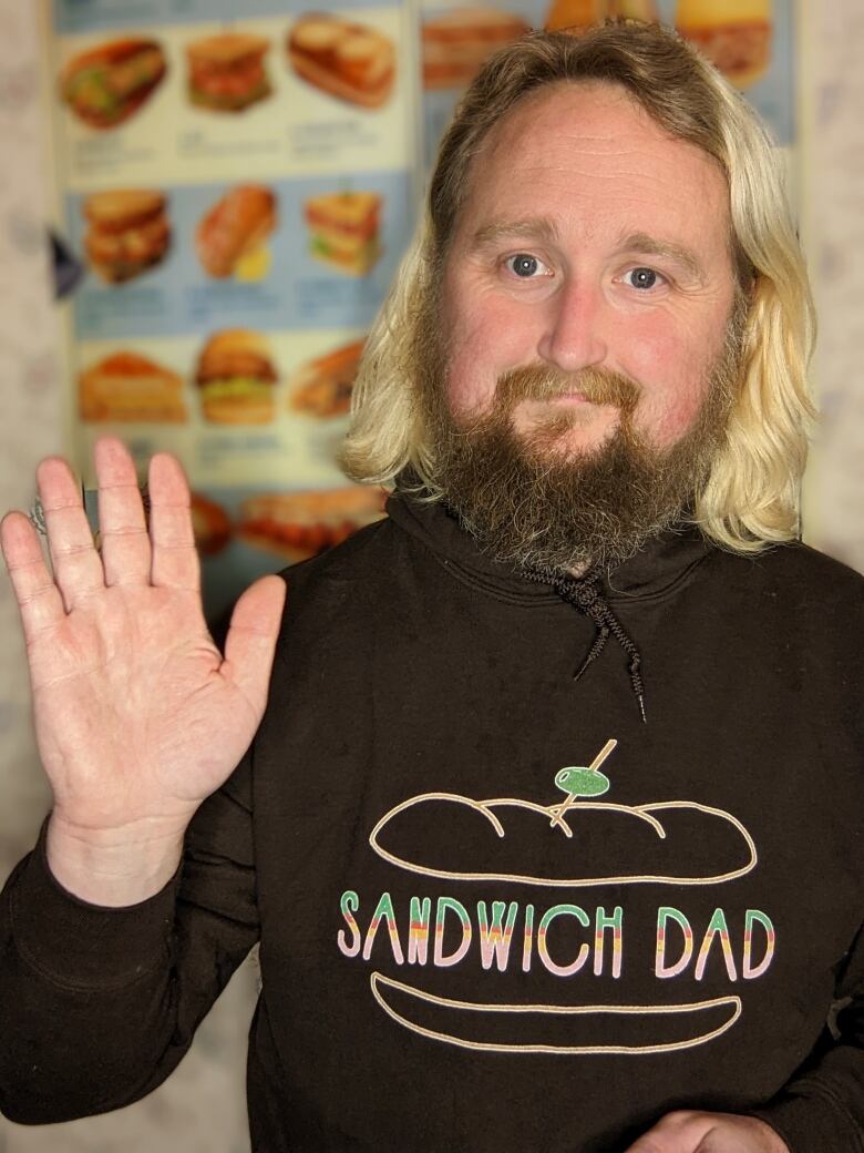 A man with blonde hair waves at the camera. He's wearing a black hoodie with the words 