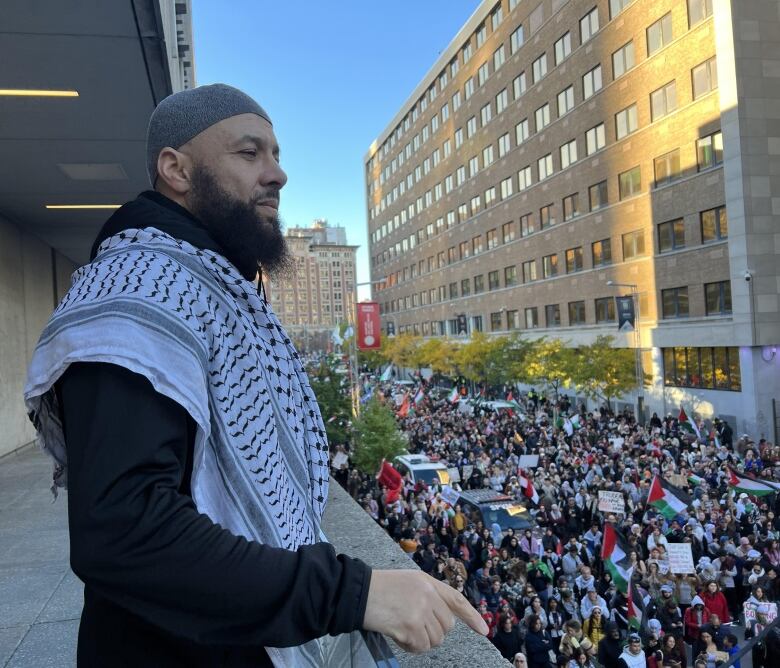 Man in a grey skullcap and a keffiyeh watches over large crowd carrying red, white and green flags.