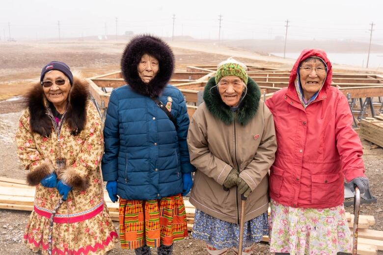 Four elderly ladies stand in a row in their jackets in front of construction. Most are smiling.