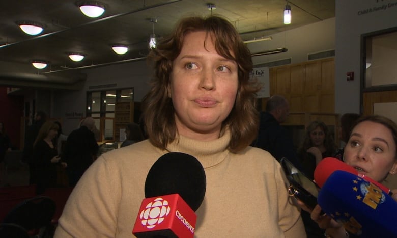 A woman in a tan shirt stands next to microphones. 