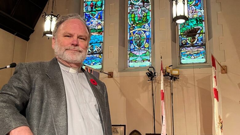 A man with grey hair and a grey beard and a priest's collar, stands in the foreground, in front of stained glass windows.