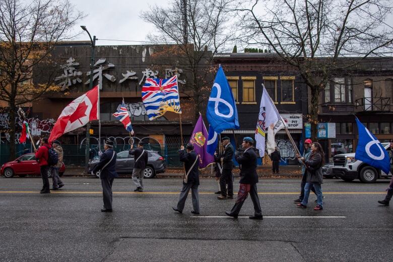 Veterans in uniforms and community members walk down the middle of a street holding various flags, including one of Canada and British Columbia. 