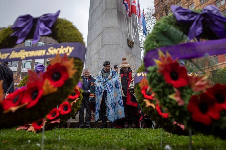 In front of a cenotaph, people are standing with their heads bowed down. They are wearing blankets and hats with Indigenous designs. In the foreground are wreaths with poppies and purple ribbons. 