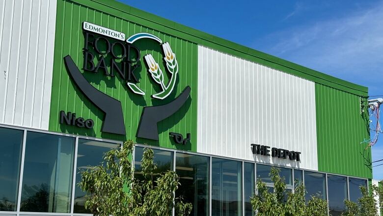 A building with a logo saying Edmonton's Food Bank. The bottom of the building is glass panels, the top is green and white panels. Some shrubs in the foreground.