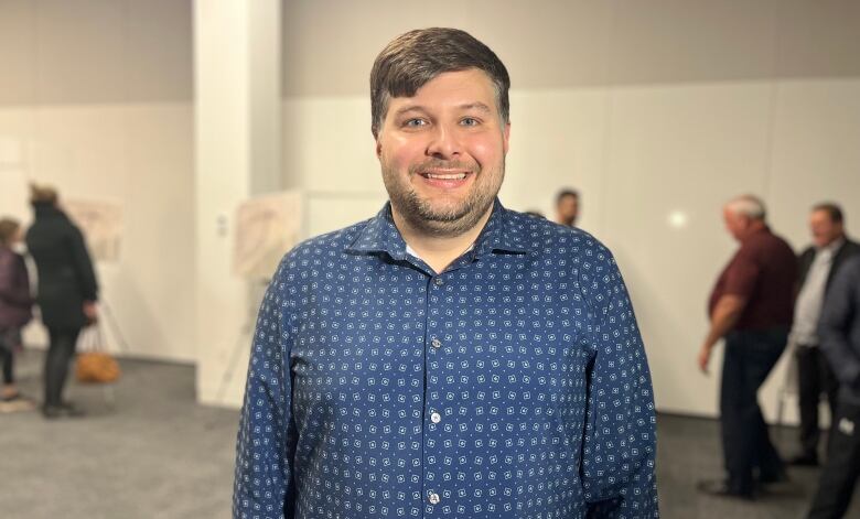 A man in a blue shirt stands in the middle of a room smiling at the camera.