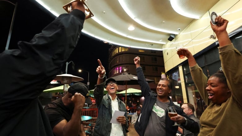 A group of five people stand in a semi-circle raising their hands in the air in celebration.