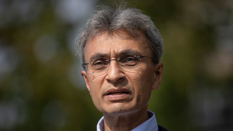 A South Asian man speaks to a mic in a park.