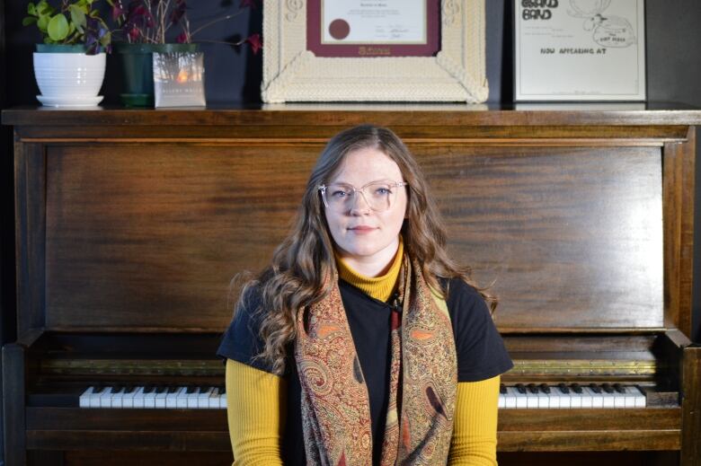 A woman with long curly brown hair and a paisley scarf sits in front of a piano. 