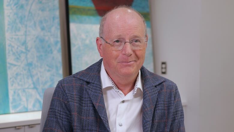 A man with un-rimmed, rectangular glasses is smiling and wearing a collared shirt with a suit.