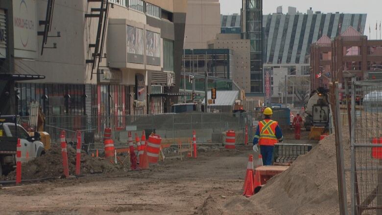 A worker in an orange safety vest walks through a torn-up stretch of road flanked by downtown buildings. 