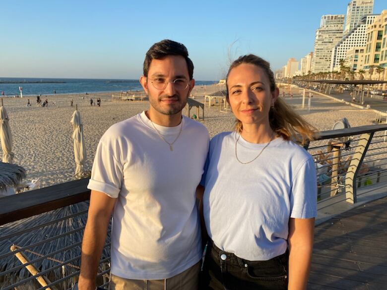 Two people smile at the beach with their arms around each other.