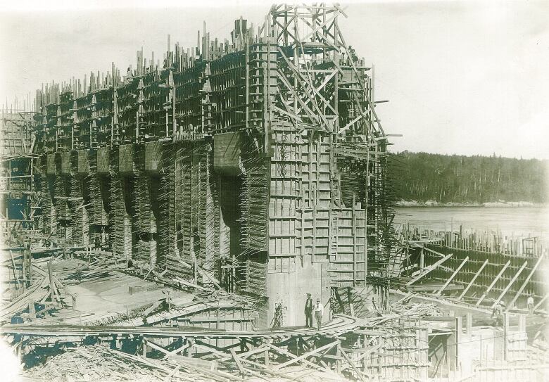 A black-and-white image of a hydro-electric dam under construction, with woods in the background