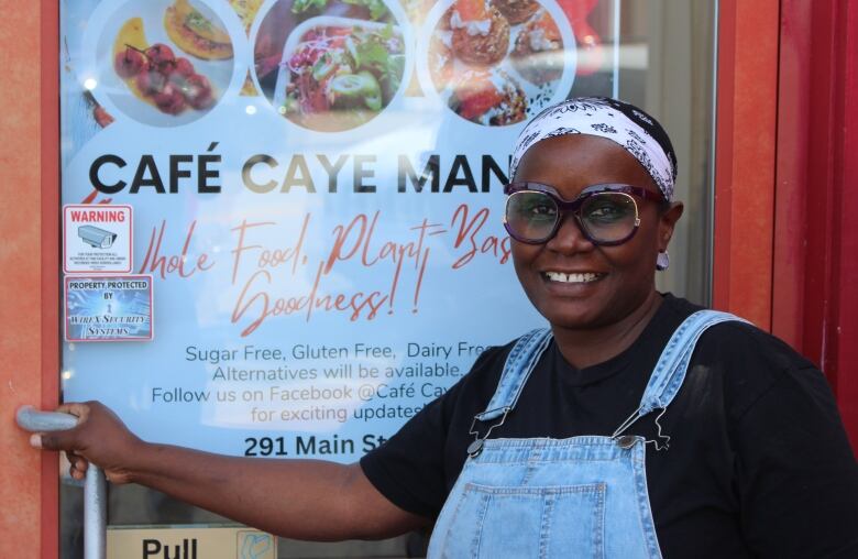 A woman in overalls and glasses stands by a glass door that bears a sign advertising a soon-to-open restaurant.