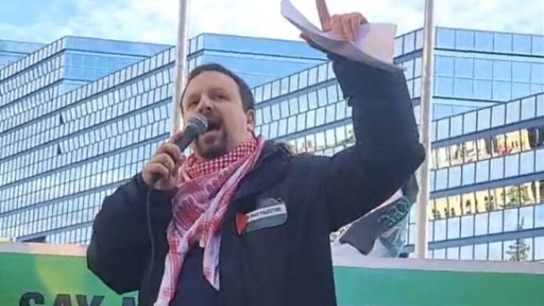 A man in a Palestinian kerchief address a crowd at a protest rally.