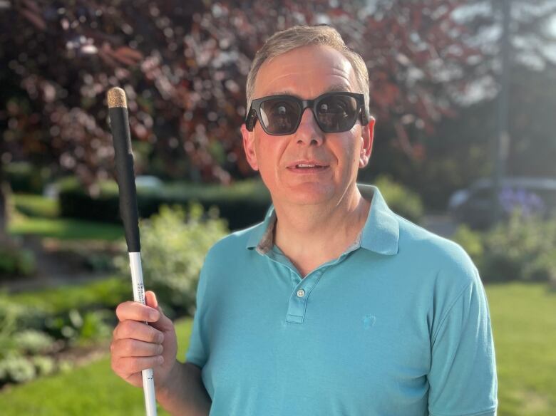 A man in a blue shirt and sunglasses holding a white cane looks into the camera. 