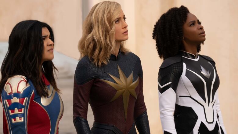 Three women stand looking up and to the left. Each wear a superhero suit. 