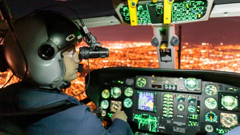 A man wearing night vision goggles flies a helicopter above the bright lights of a city.