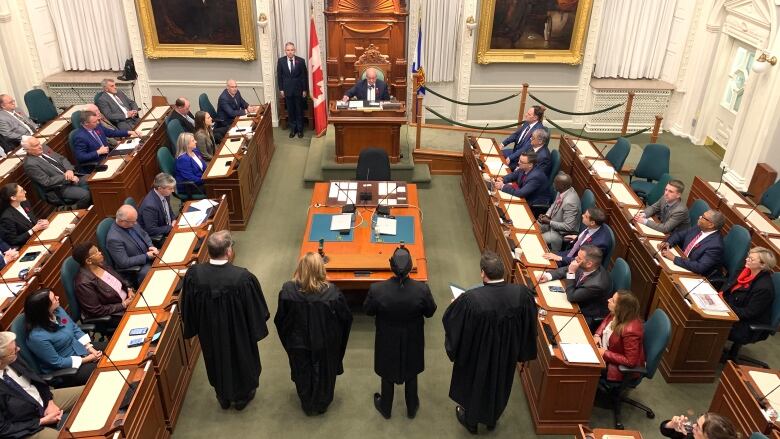 For people stand in front of a table with rows of people seated on either side.