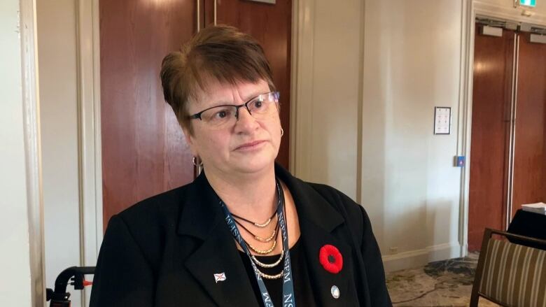 A white woman with glasses and short brown hair wears a black jacket with a red poppy on her chest. She is standing in a hotel ballroom with doors behind her