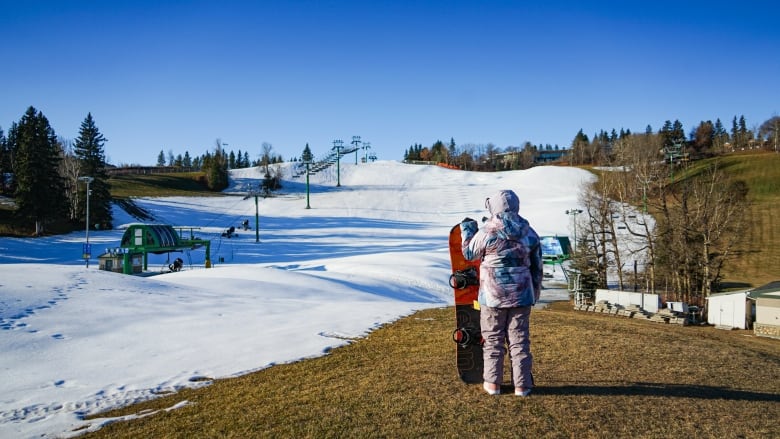 A lonely snowboarder looks over Snow Valley Ski Club on November 9, 2023. The hill is half covered with snow, with a lot of grass on the right side. 