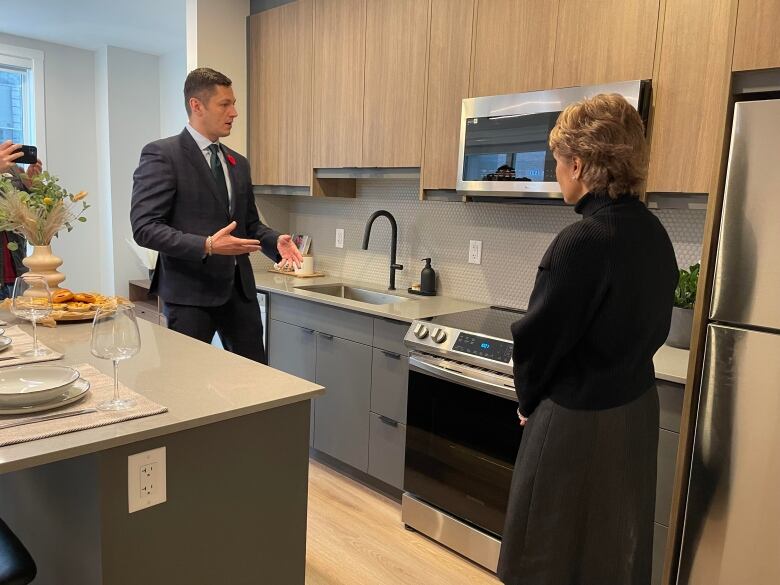 a man and woman stand in a kitchen speaking to each other