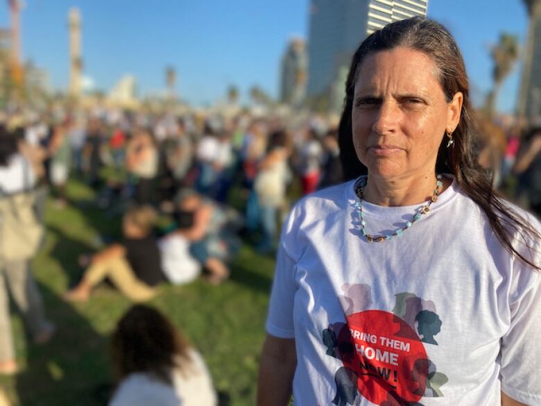 A woman with long, brown hair wearing a t-shirt with the slogan 'bring them home now' stands with a crowd of people in a city park. 