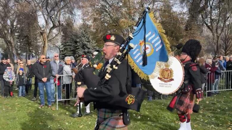 Two bagpipers and a drummer in ceremonial military uniforms march and play as a crowd wearing red poppies watches.