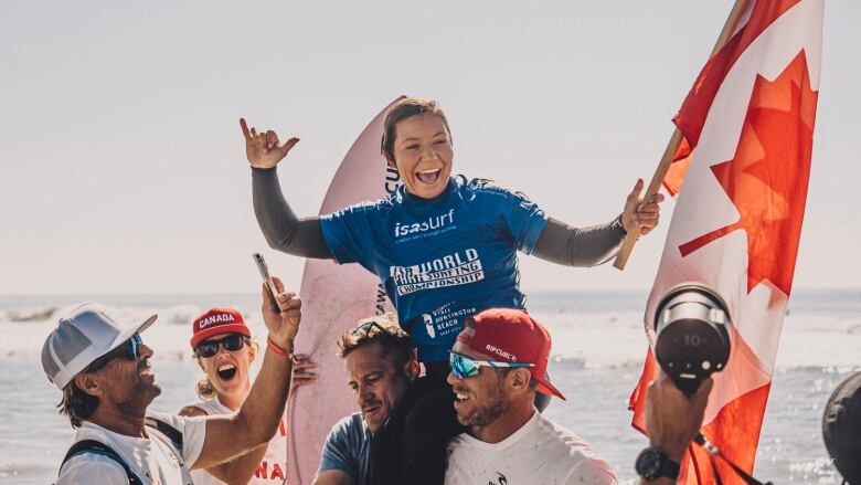 A female Para surfer smiles while holding a Canadian flag in her left hand as coaches hoist her up on their shoulders following a competition.