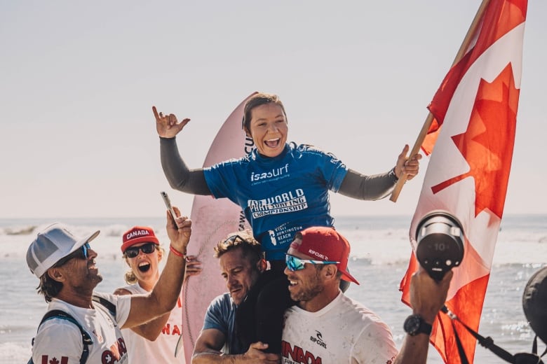 A female Para surfer smiles while holding a Canadian flag in her left hand as coaches hoist her up on their shoulders following a competition.