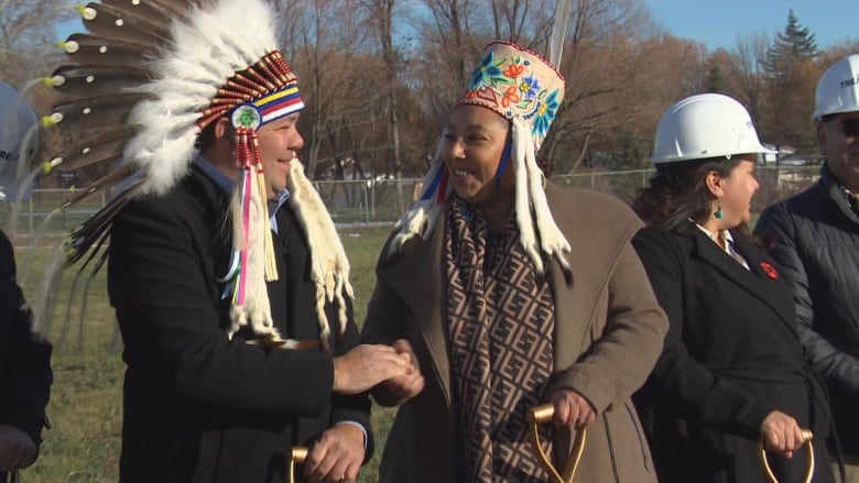 A man and a woman wearing headdresses shake hands while holding shovels.