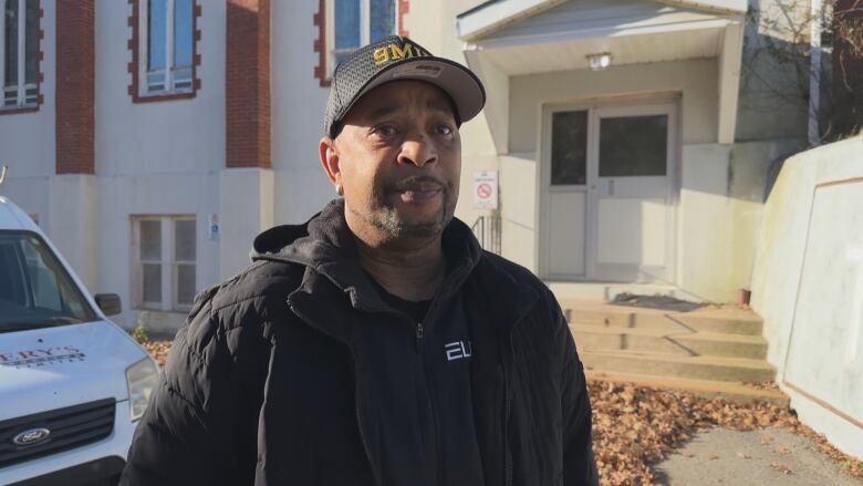 A man stands in front of a church. He is wearing a black hoodie, black jacket, and black ball cap.