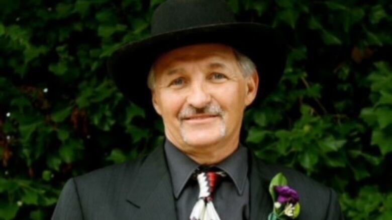 A man wearing a black suit, black hat and a red tie is pictured standing with a wine glass in front of some trees.