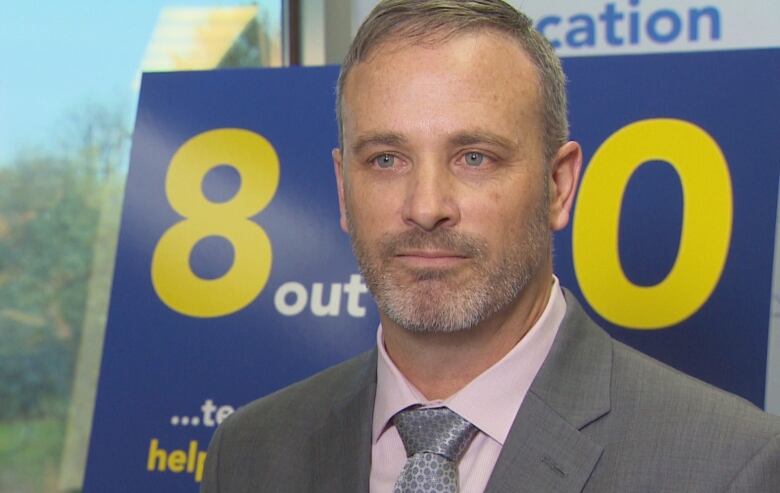 A man with light hair and a short beard. He is wearing a grey suit and pink dress shirt.