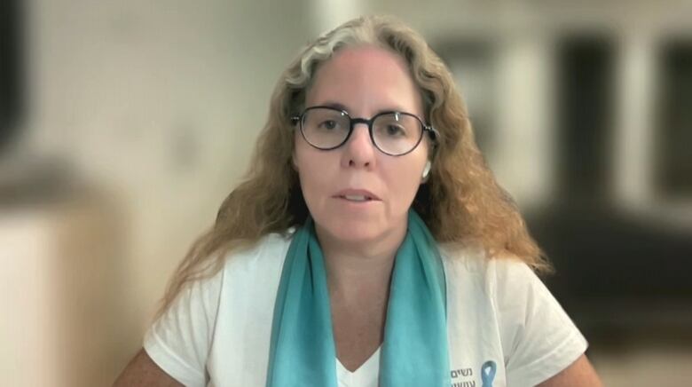 A woman with long, wavy hair and glasses looks at a camera.