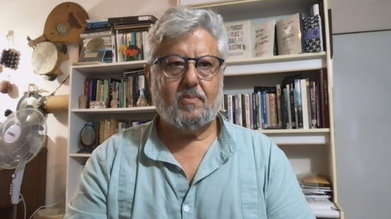 A man in white, wavy hair, a white beard and glasses looks at the camera. He wears a light blue shirt. A bookcase stands behind him.