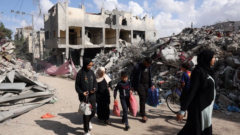 People carry belongings as they walk past the rubble of destroyed buildings.