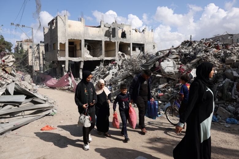 People carry belongings as they walk past the rubble of destroyed buildings.