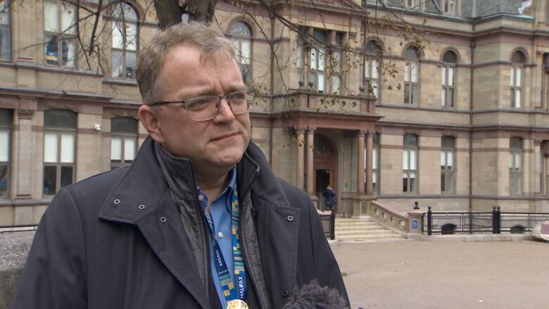 A white man with glasses wears a black jacket and blue collared shirt. He stands outside City Hall, which is a tall beige stone building