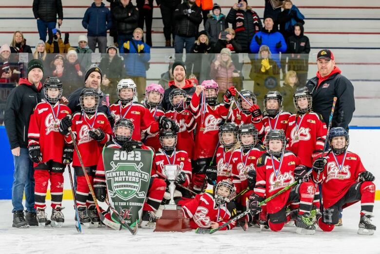 A group of young hockey players in red, white and black uniforms and black helmets pose for a group picture, with a championship banner and trophy in front of them.