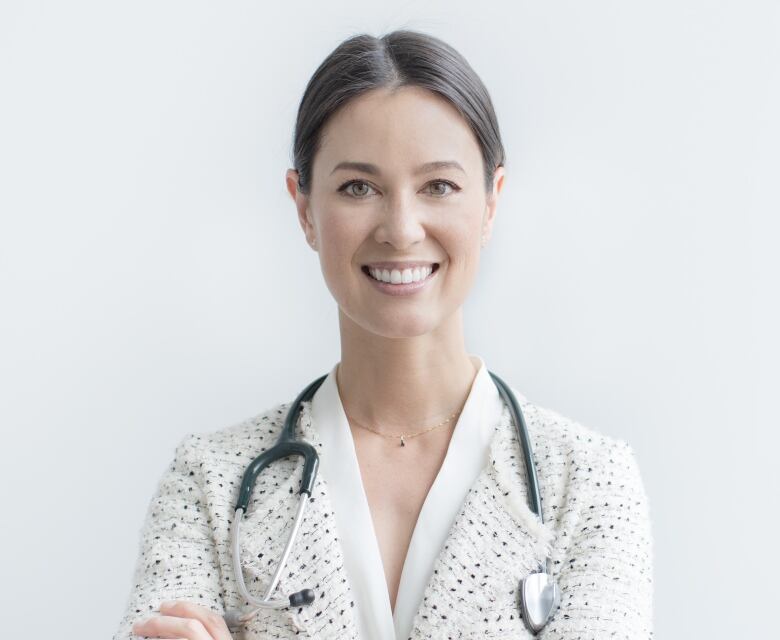 A woman wearing a blazer and stethoscope smiles while posing for a photograph with her arms crossed.