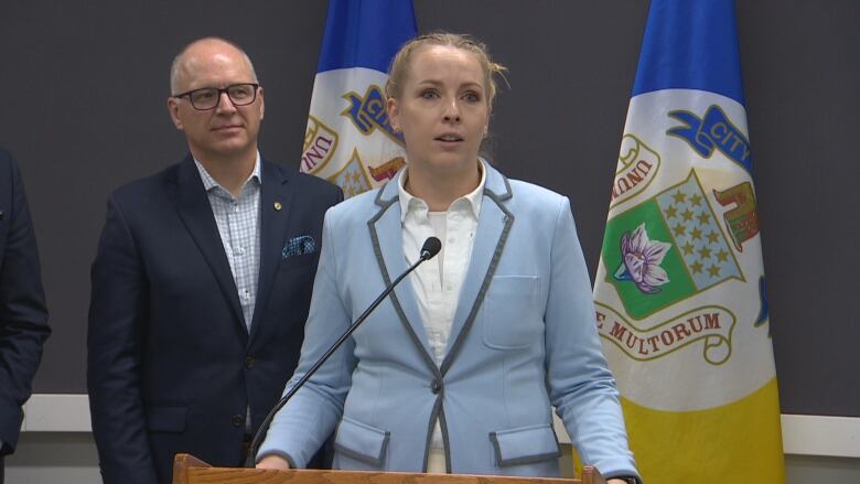 A woman in a light blue blazer is speaking at a modium, while a man in a black blazer, standing behind, looks on.