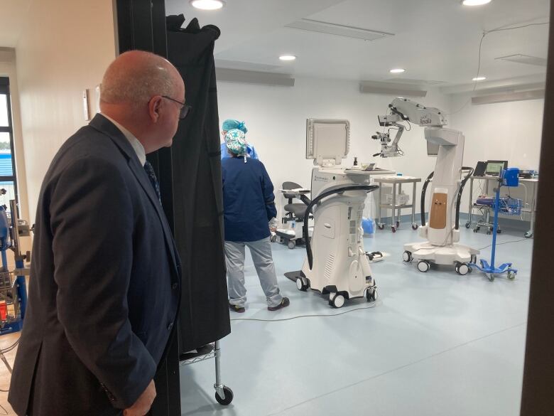 Man with silver hair and glasses wearing suit jacket and tie looks into room where there is medical equipment set up.