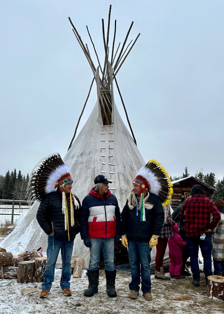 Two men in headdresses look at a man between them in front of a teepee.