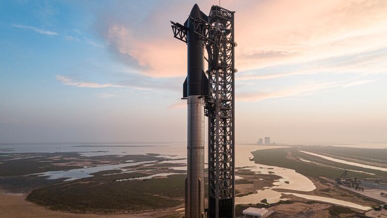 A silver and black rocket is seen sitting on a launch pad near the ocean.